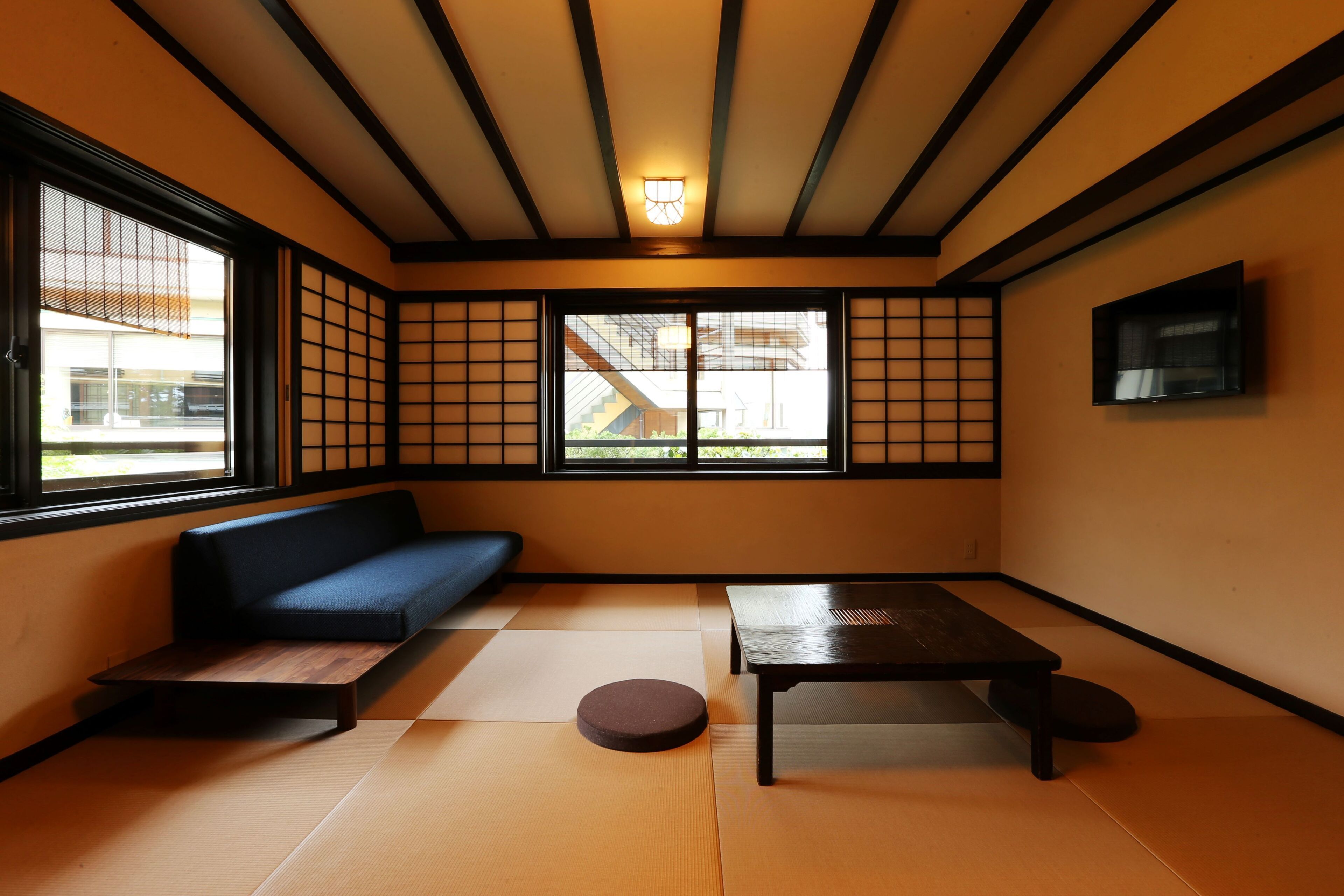 A traditional Japanese room with tatami flooring, a low table, a sofa, and a television mounted on the wall.