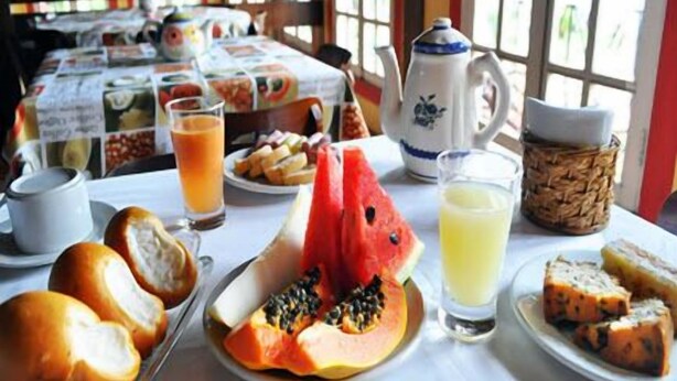 A breakfast setting with a table set for two, featuring a plate of watermelon, papaya, and bread, along with a pitcher of juice and a teapot. - Vale Verde Praia Hotel (Porto Seguro)