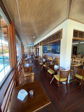 A spacious restaurant with wooden tables and chairs, large windows, and a blue wall with kitchen equipment. - Hotel Porto das Âncoras (Porto Seguro)