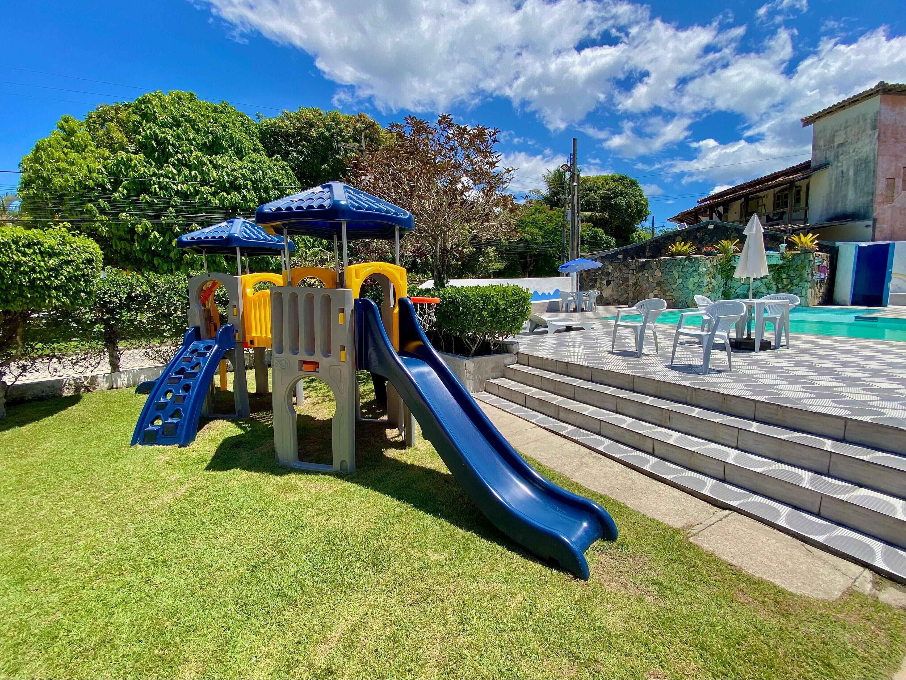 A playground with slides and climbing structures, a pool area with lounge chairs, and a stone wall with greenery.