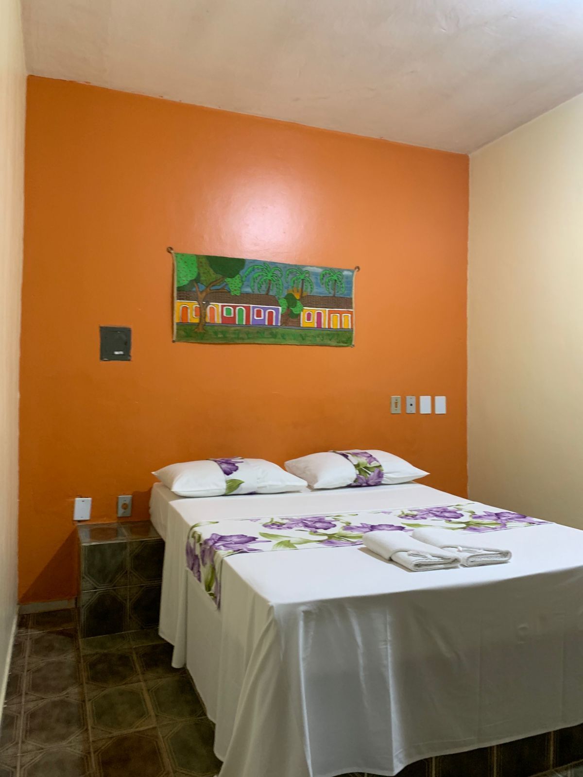 A single bed with a white bedspread and purple floral pillows in a room with orange walls.