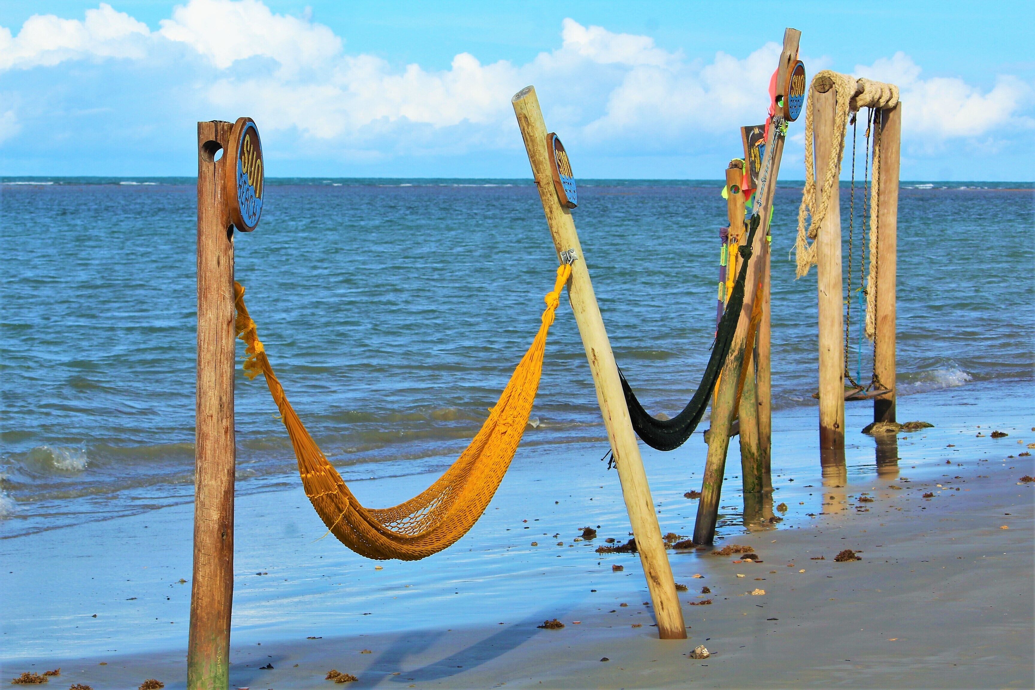 Un hamac est tendu entre deux poteaux en bois sur une plage, avec l’océan en arrière-plan.