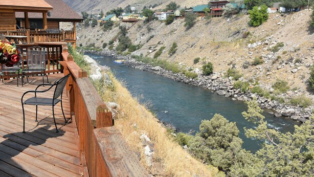 A wooden deck overlooking a river with a view of houses and mountains in the background. - Yellowstone River Suites (Gardiner)