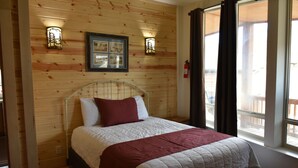 A wooden-paneled bedroom with a bed, two wall-mounted lights, a framed picture, and a window with curtains. - Yellowstone River Suites (Gardiner)