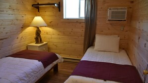 Una habitación tipo cabaña de madera con dos camas, una mesita de noche con lámpara, una ventana con cortinas y un equipo de aire acondicionado.