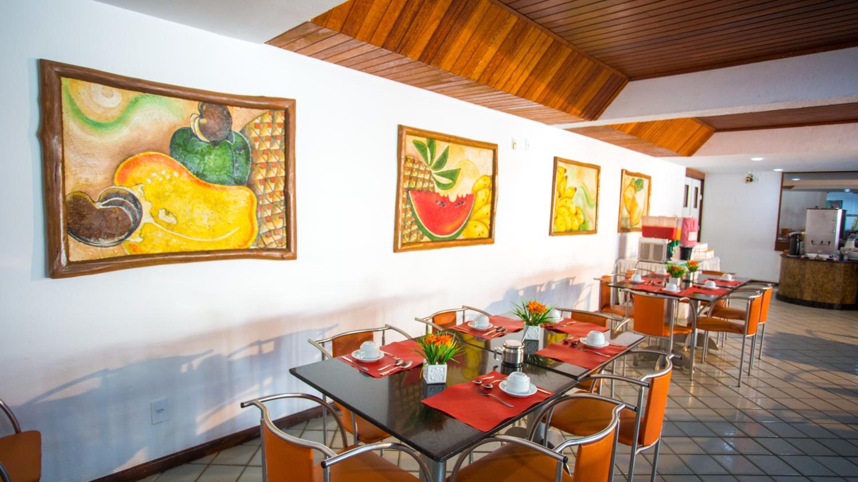 A dining area with tables and chairs, artwork on the walls, and a kitchen in the background.