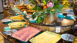 A buffet with various dishes, including meats, cheeses, and desserts, arranged on a counter with a decorative plant. - Monte Pascoal Praia Hotel (Porto Seguro)