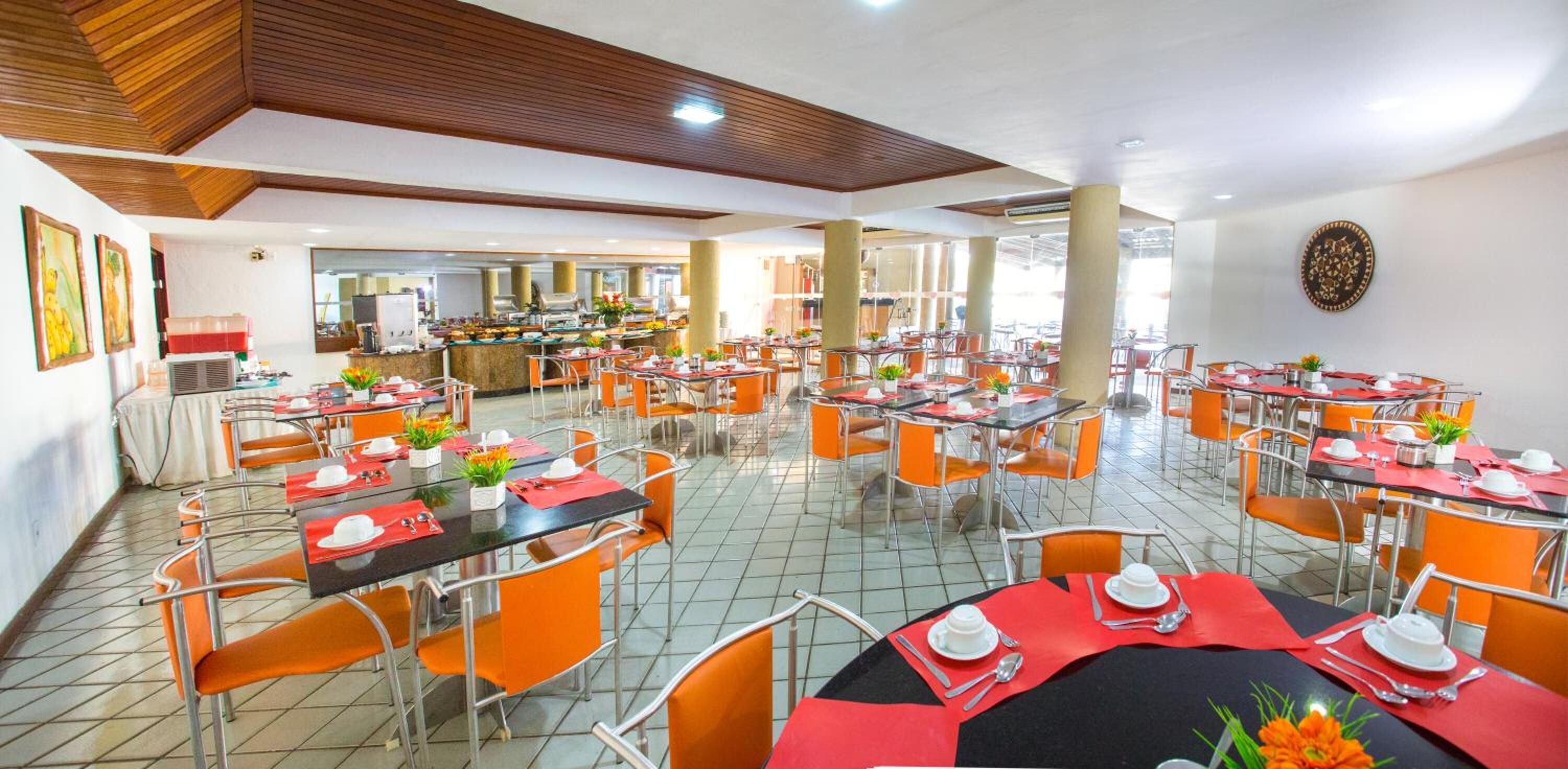 A spacious dining area with multiple tables and orange chairs, featuring a central buffet and a large mirror on the back wall.