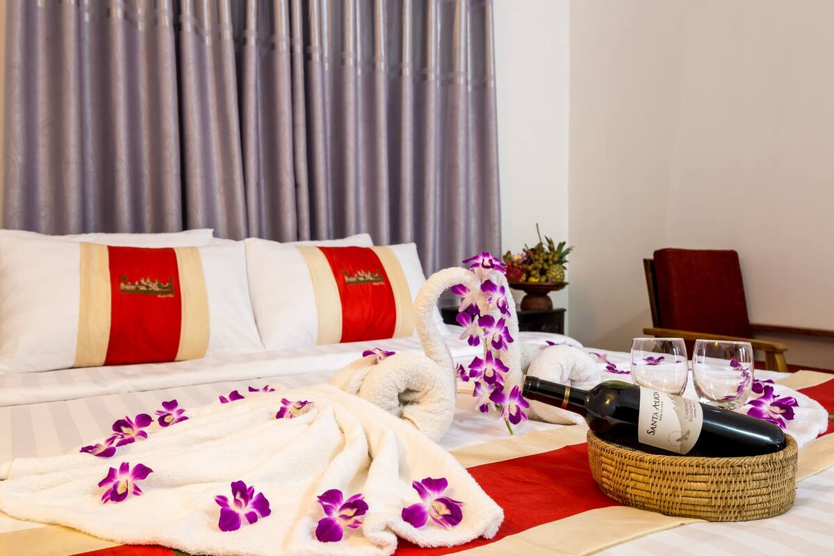 A hotel room with a neatly made bed, a bottle of wine, two glasses, and a decorative orchid arrangement.