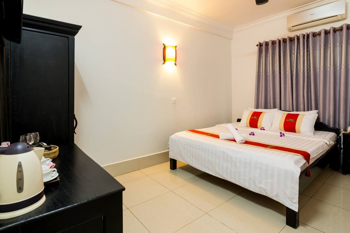 A hotel room with a bed, a nightstand, a desk with a kettle, and a wall-mounted light.