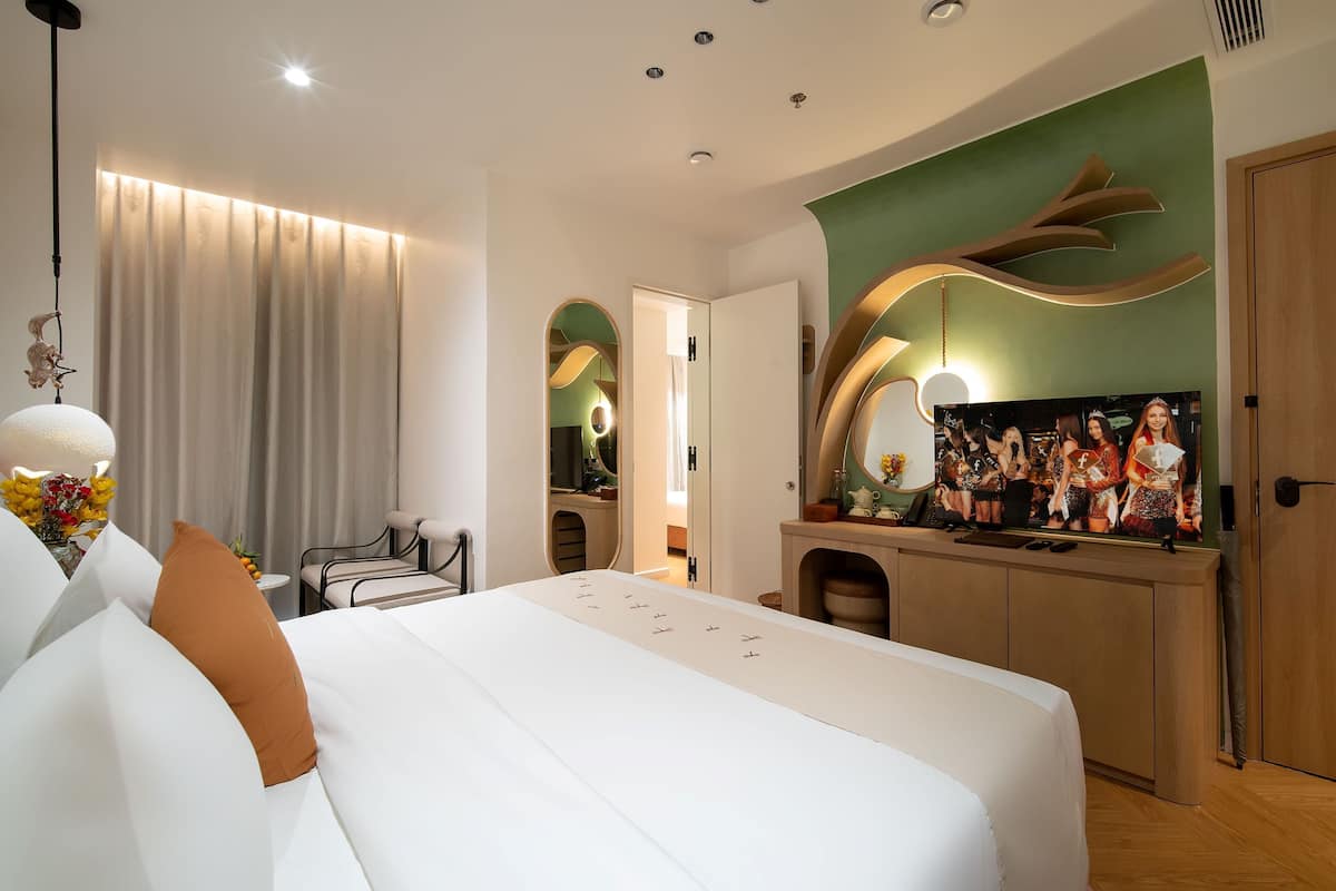 A modern hotel room with a large bed, a vanity with a mirror, and a television displaying a group photo.