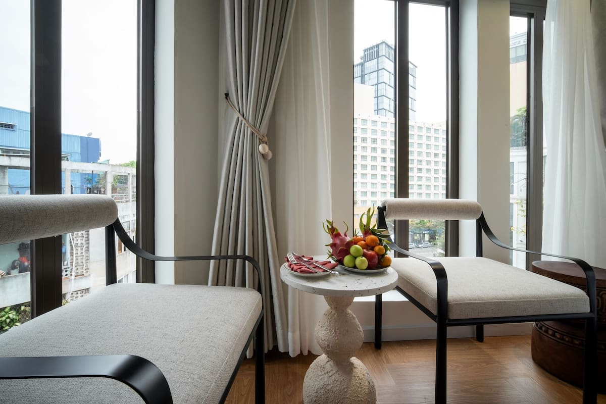 A modern hotel room with two chairs, a small round table with fruit, and a view of the city through large windows.
