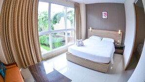 A hotel room with a large bed, a nightstand, a bedside table, and a view of greenery outside. - Tamnak Villa (Pattaya)