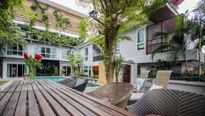 A poolside area with wicker chairs and a wooden table, surrounded by a building with balconies and greenery. - Tamnak Villa (Pattaya)