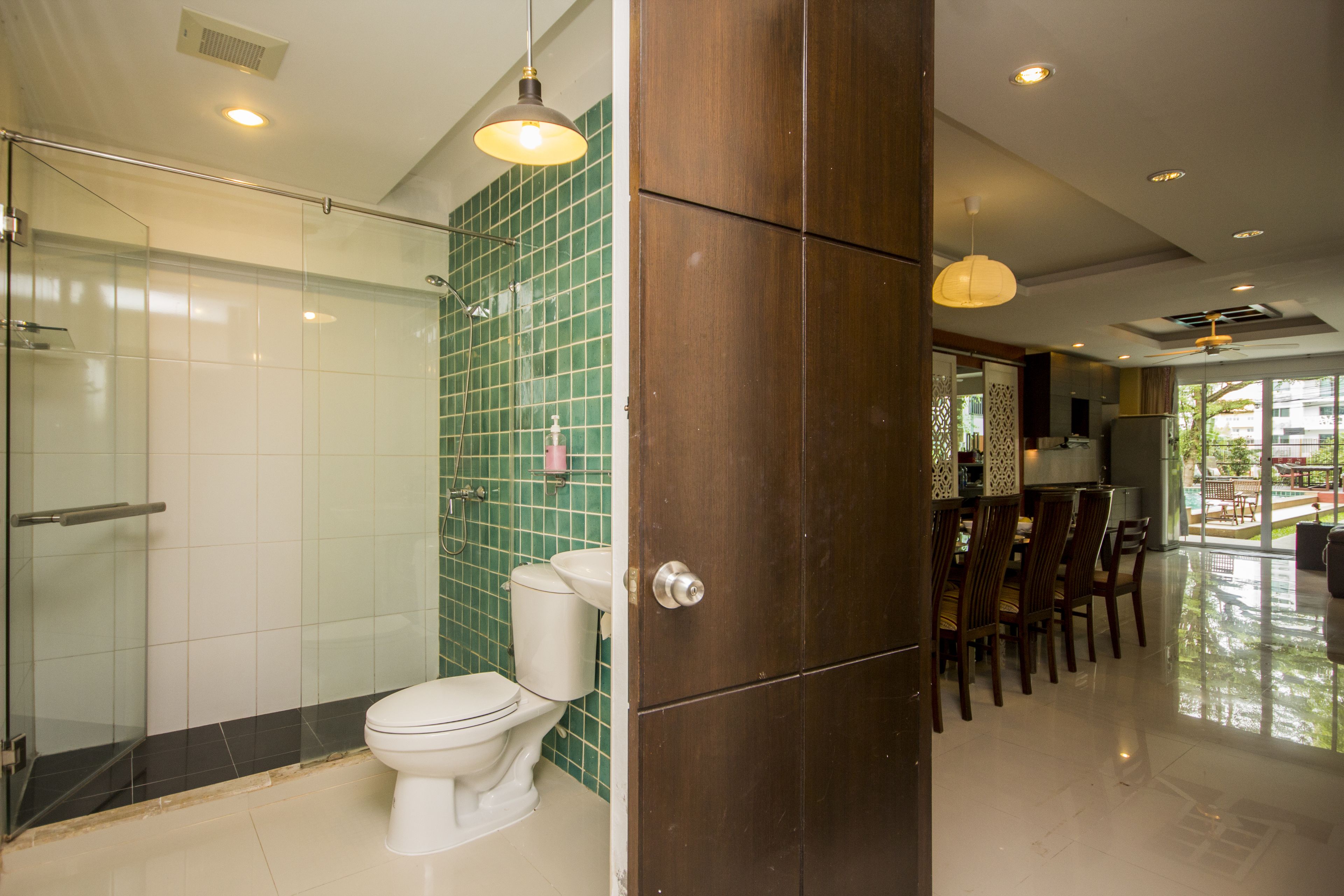 A modern bathroom with a glass shower enclosure, a toilet, and a wooden door leading to a dining area with a view of the outdoors.