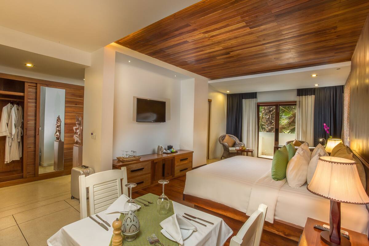 A hotel room with a wooden ceiling, a bed, a desk with a chair, a TV, and a window with curtains.