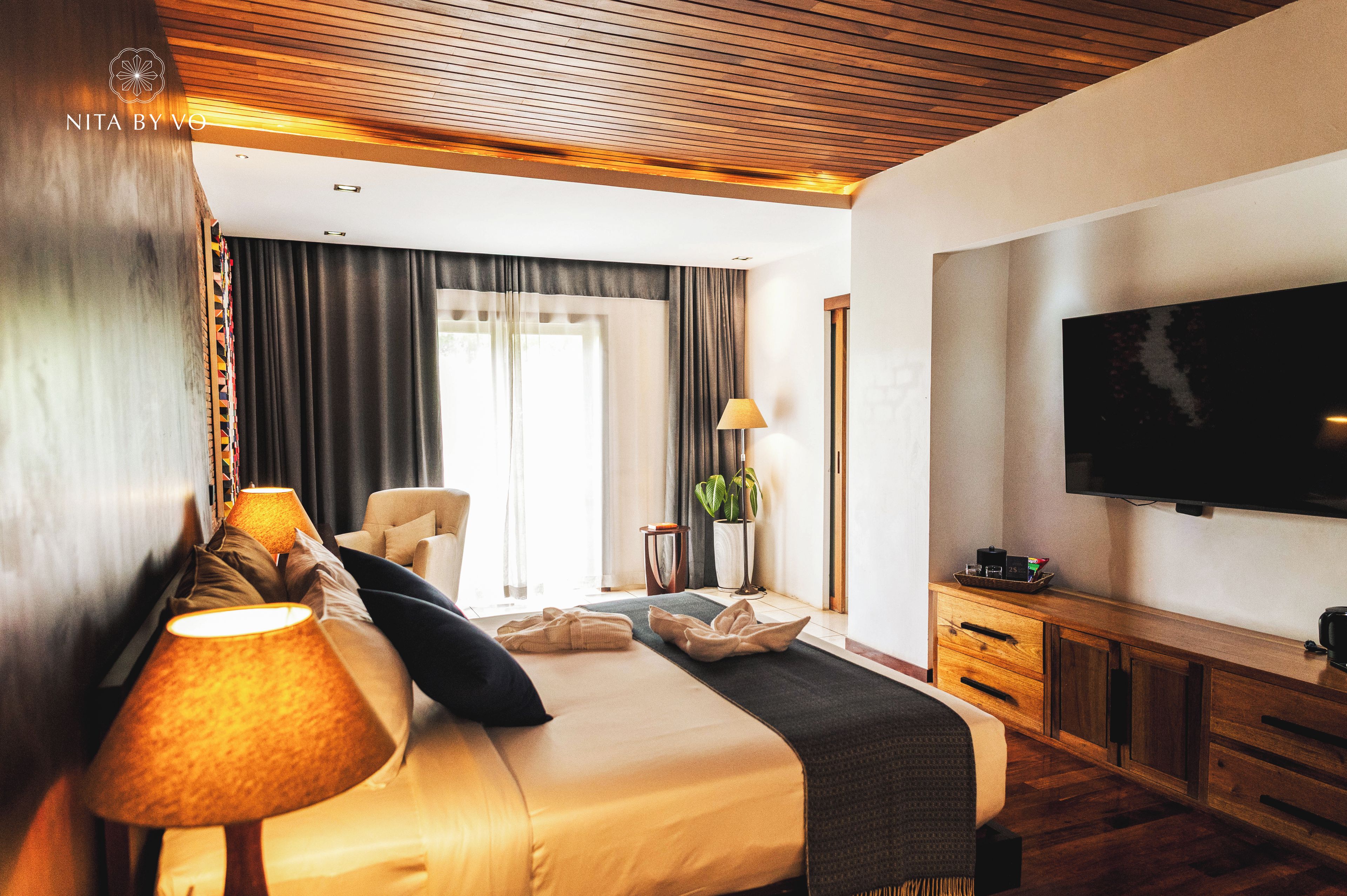 A hotel room with a bed, a TV, a desk, and a large window with curtains.