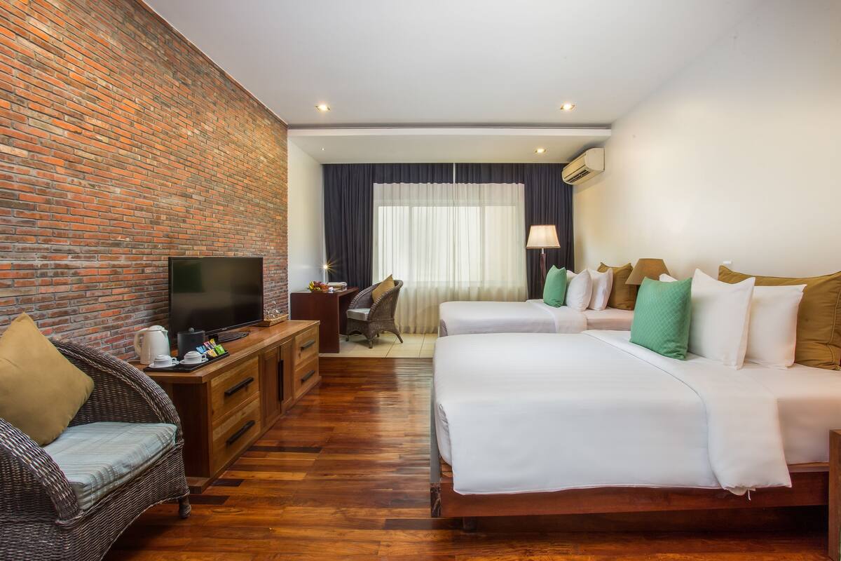 A hotel room with a brick wall, a flat-screen TV, a wooden dresser, a wicker sofa, two beds with white bedding, and a desk with a chair.