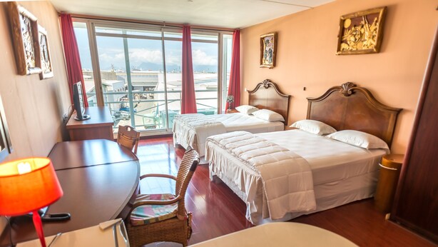 A hotel room with two beds, a desk, a chair, a window with a view of the city, and a painting on the wall. - Hotel Sarah Nui (Papeete)