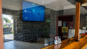 A reception area with a wooden reception desk, a mounted television displaying the time, and a stone wall. - Hotel Sarah Nui (Papeete)