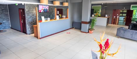A modern hotel lobby with a reception desk, a TV mounted on the wall, and a seating area with a plant and a vase of flowers.