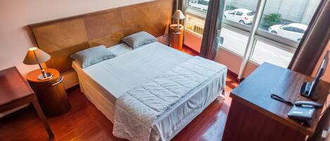 A hotel room with a wooden headboard, a bed with a white comforter, a nightstand with a lamp, a desk with a telephone, and a window with a view of parked cars.