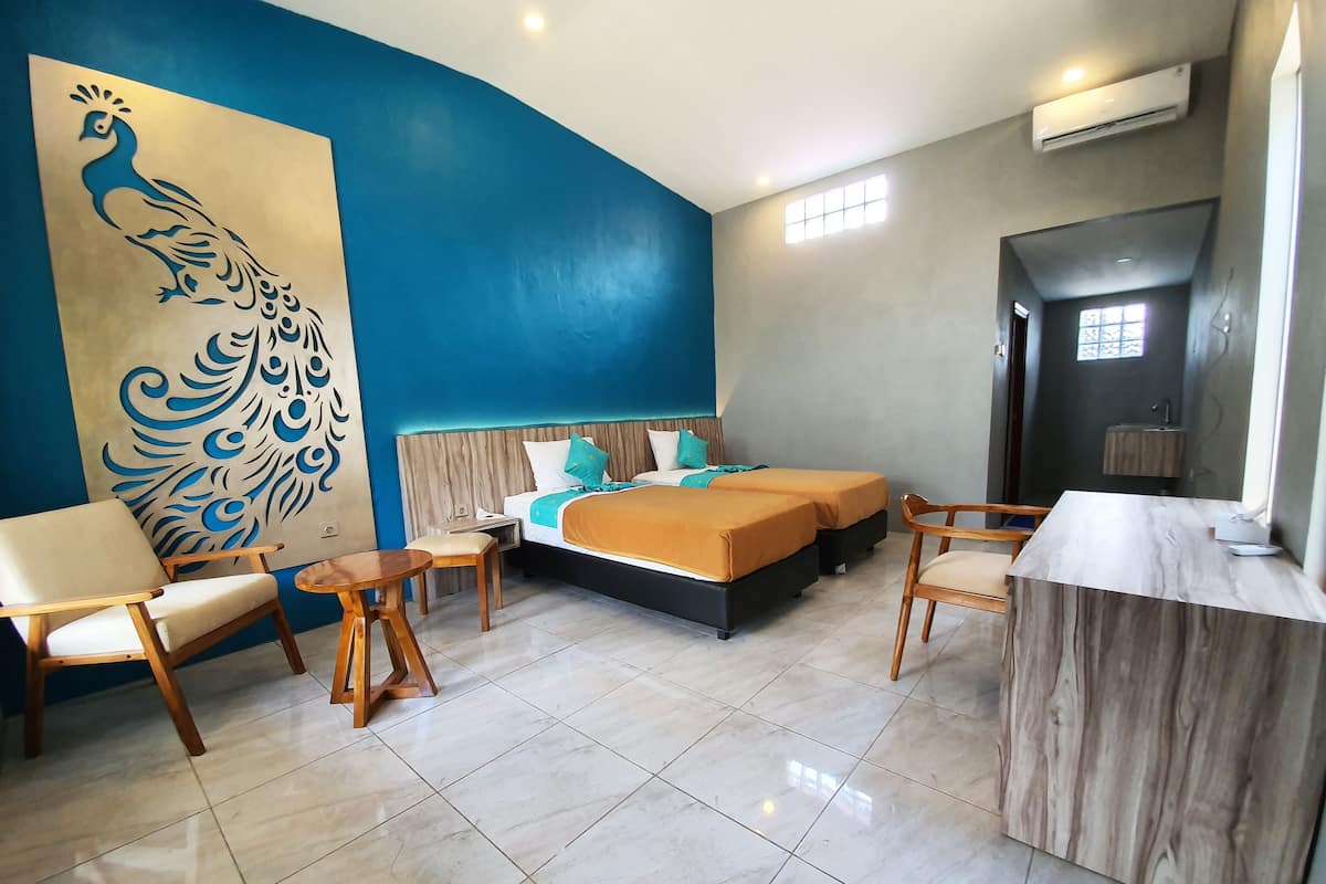 A hotel room with a large peacock mural, a bed with orange bedding, two bedside tables, and a wooden chair.