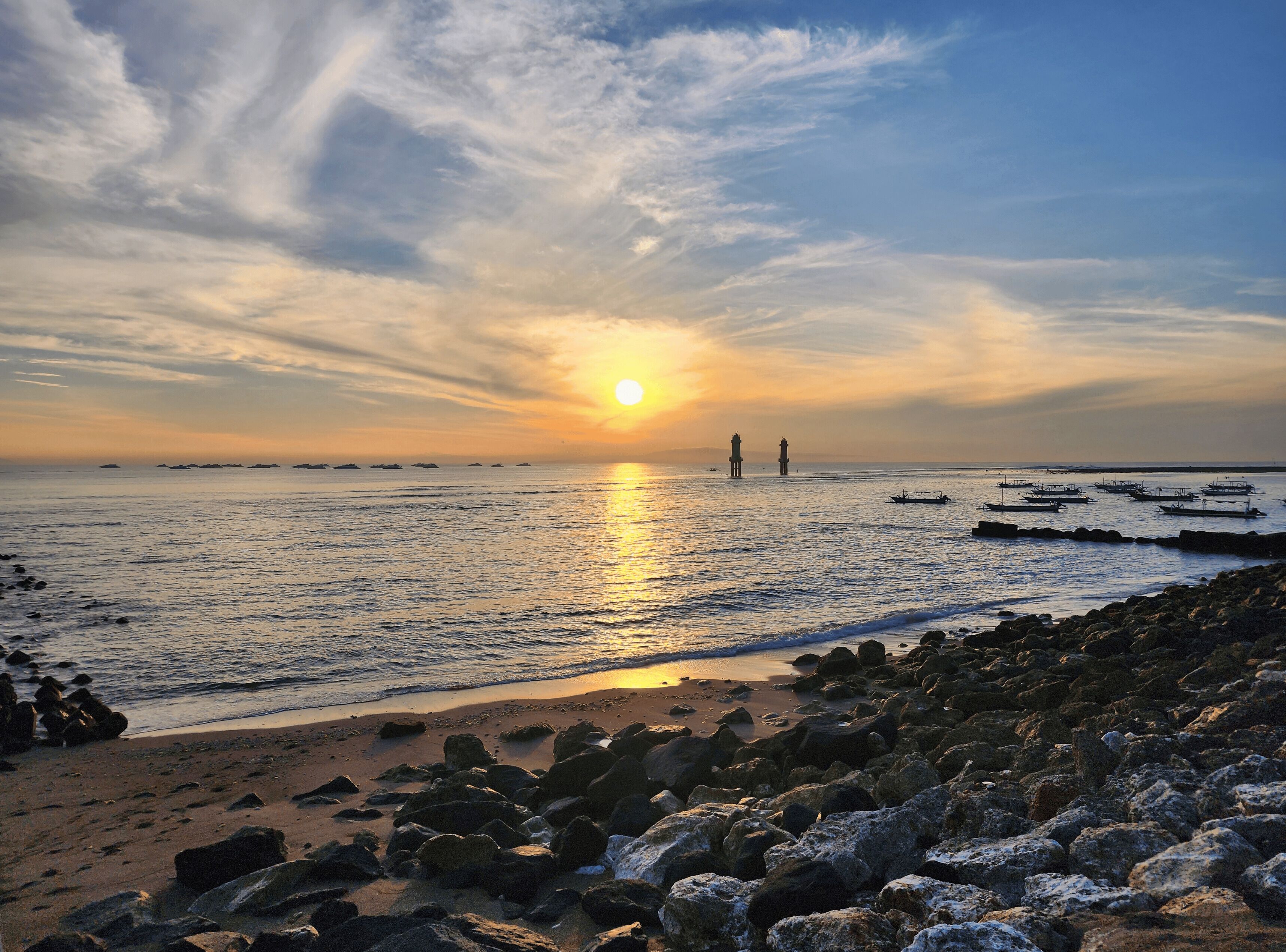 Une plage avec des rochers au premier plan, une mer calme et deux phares à l’horizon.