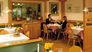 En hyggelig restaurant med træmøbler, borde dækket med stearinlys og blomster, samt et vindue med planter.