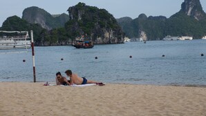 A beach with two people lying on towels, a volleyball net, and boats in the background. - Syrena Cruises (Tuan Chau)