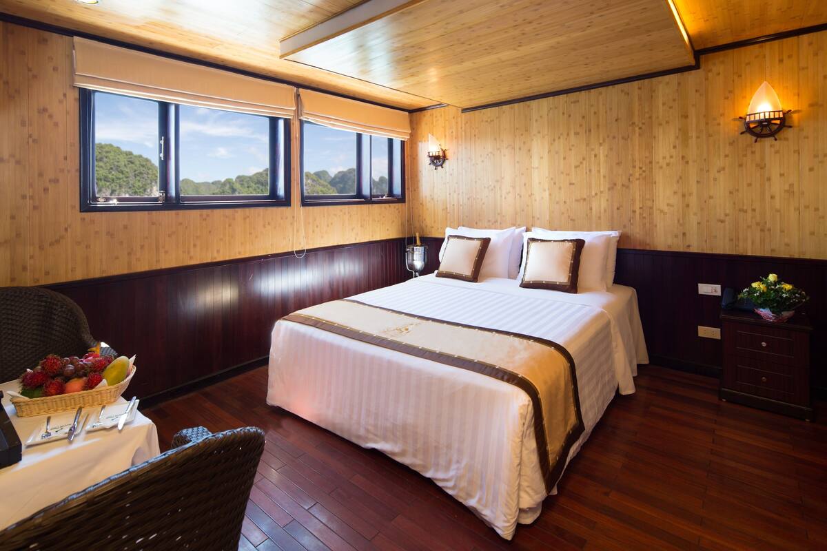 A hotel room with a large bed, wooden panel walls, a window with a view, a bedside table with a fruit basket, and a nightstand with a lamp.