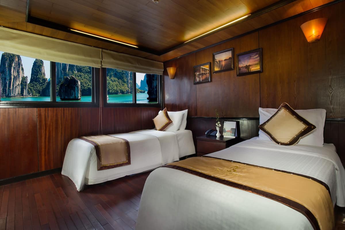 A hotel room with two beds, wooden paneling, large windows with a view of a mountainous landscape, and a nightstand with a lamp.