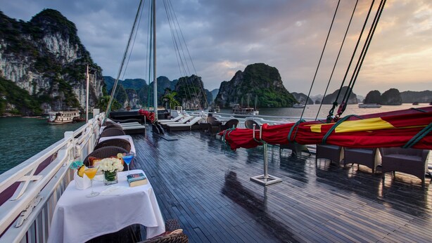 A boat deck with tables set for dining, a view of the water and surrounding cliffs, and a rope barrier. - Syrena Cruises (Tuan Chau)