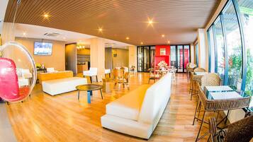 A spacious lobby with a wooden floor, white sofas, wicker chairs, and a large window offering a view of the outdoors.