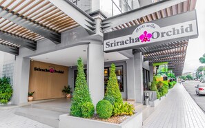 Exterior de un hotel llamado 'Sriracha Orchid' con un diseño arquitectónico moderno, que presenta una entrada destacada, plantas en macetas y un corredor cubierto.