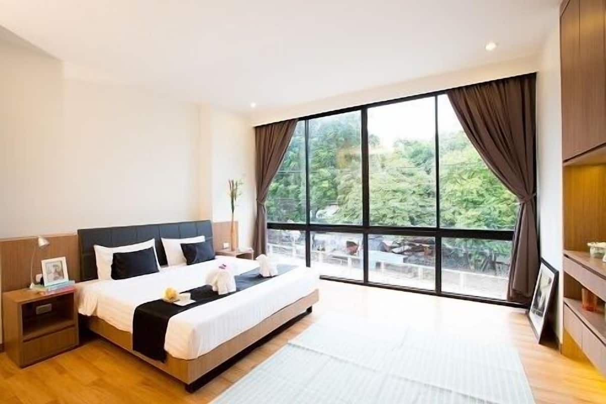 A modern hotel room with a large bed, a nightstand, a bedside table, and a view of greenery through large windows.