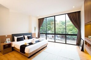 Una habitación de hotel moderna con una cama grande, una mesita de noche y vistas a zonas verdes a través de amplias ventanas.