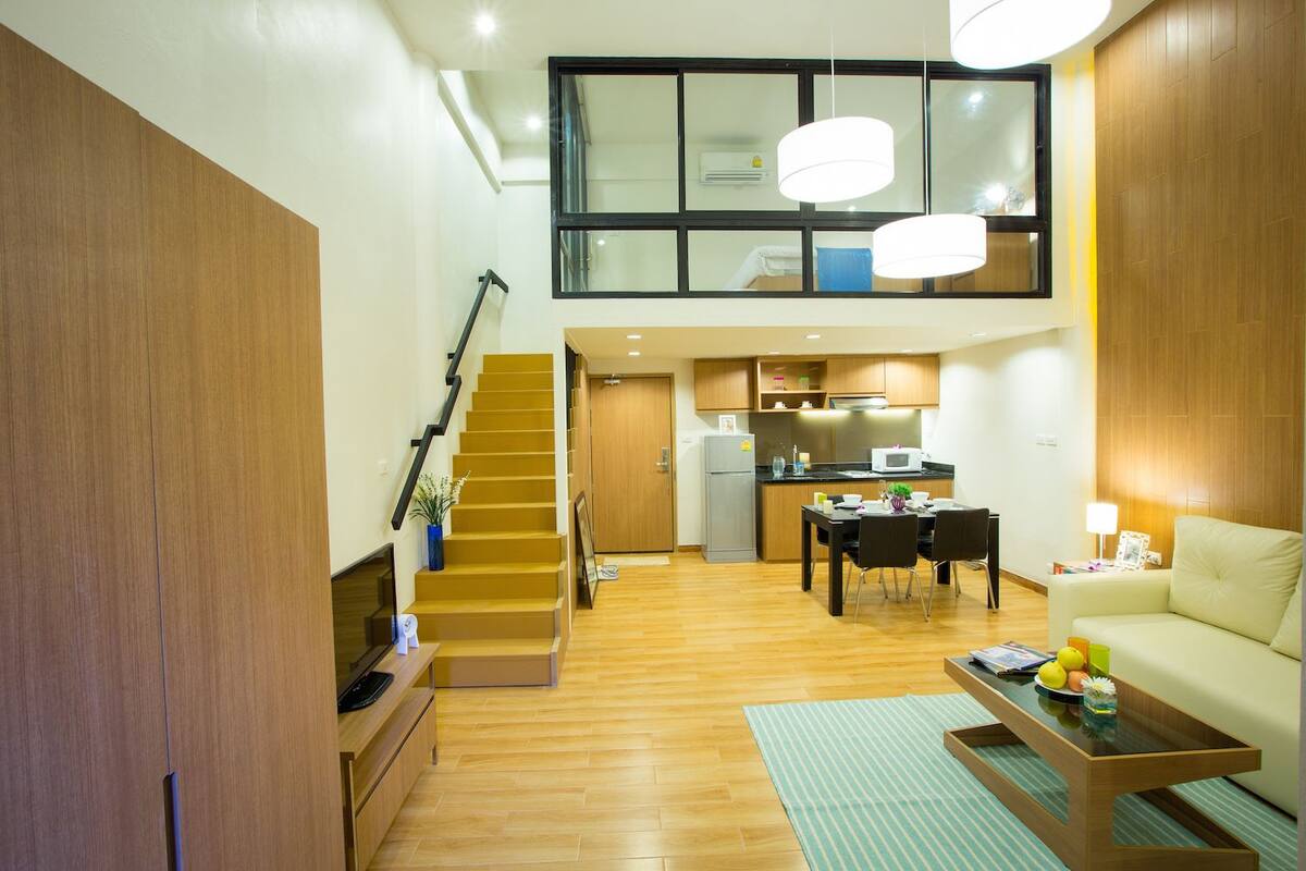 A modern apartment with a wooden staircase, a dining area, a kitchen, and a living room with a sofa and coffee table.