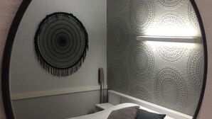 A circular mirror reflecting a bed with two pillows, a wall with a circular pattern, and a light fixture. - L'Acanthe Hôtel (Erbree)