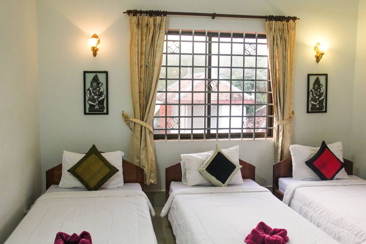 A hotel room with three single beds, each with a pillow and a towel. There are two wall-mounted lights and a window with a metal grid.