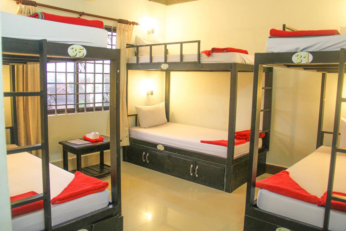 A dormitory room with bunk beds, a small table with a lamp, and a window with curtains.