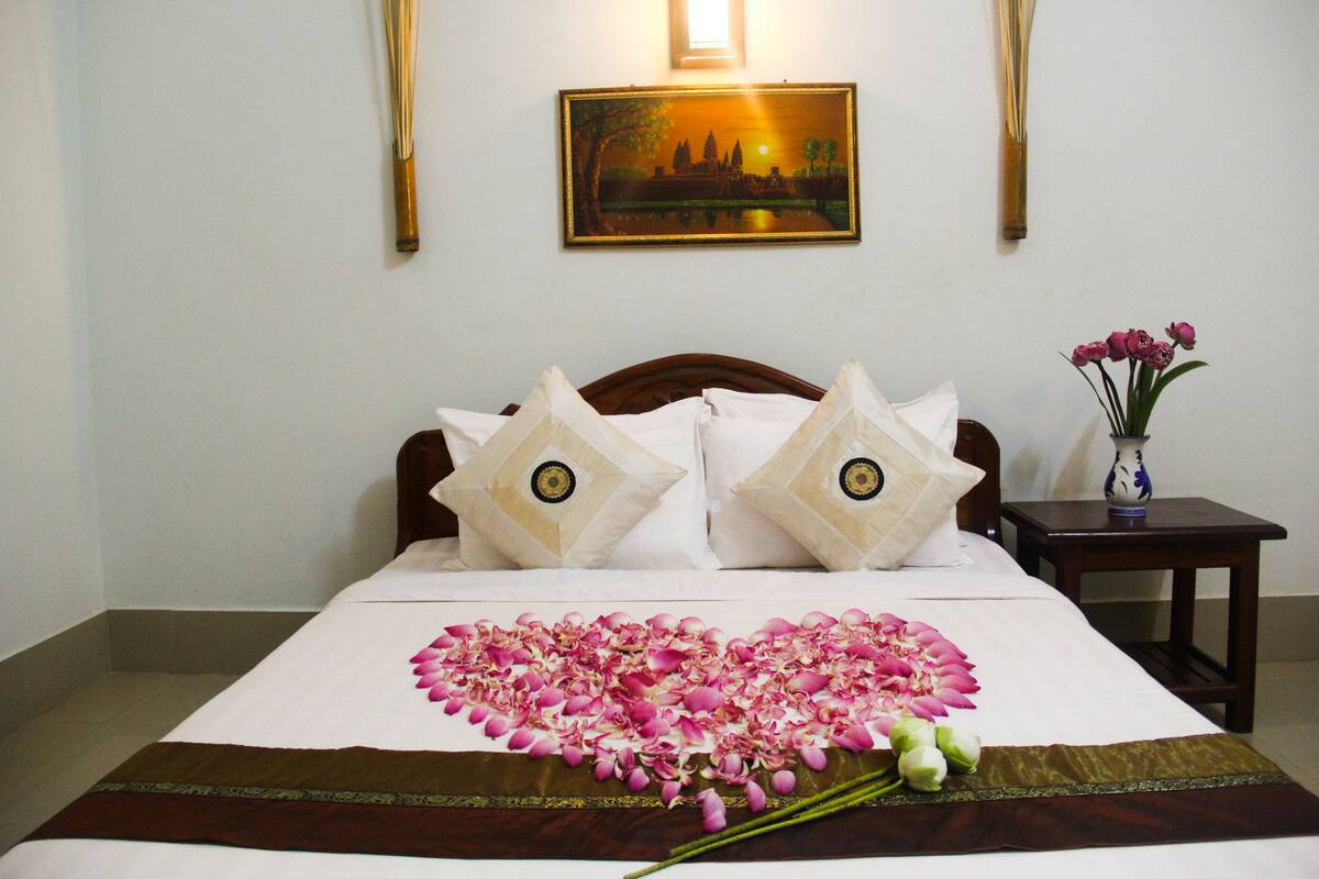 A neatly made bed with a heart-shaped flower arrangement and a vase of flowers on a nightstand.