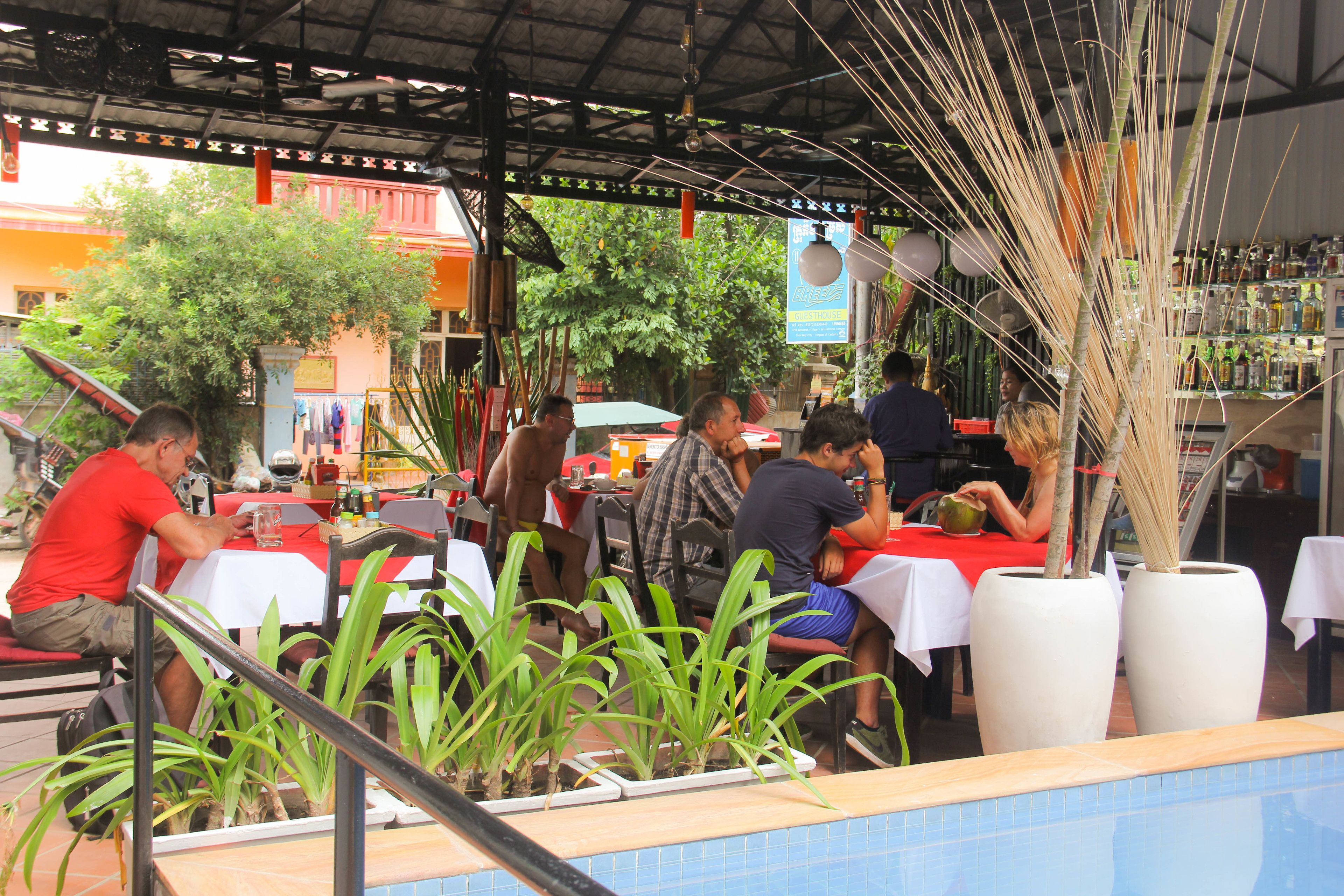 An outdoor restaurant with tables and chairs, surrounded by potted plants and a pool.