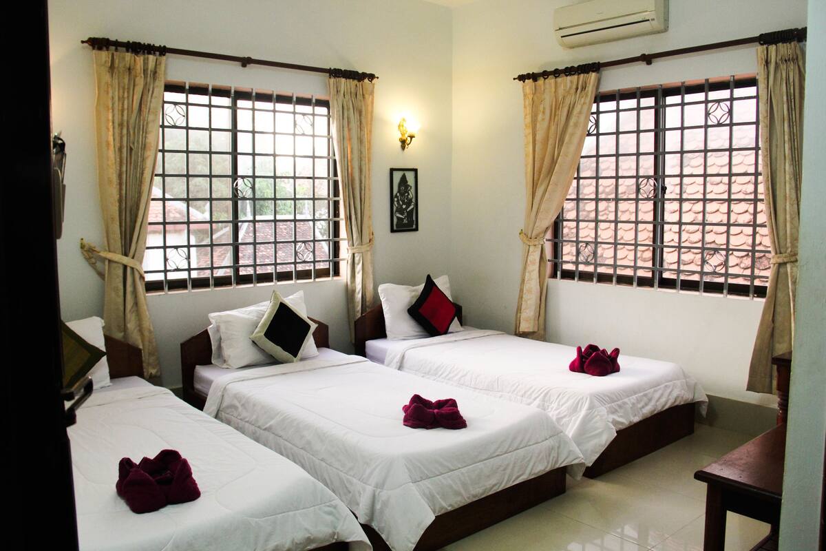 A hotel room with three beds, each with white bedding and red towels. There are curtains on the windows, and a wall-mounted air conditioner is visible.