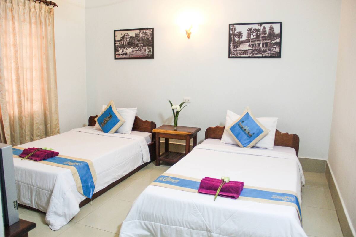 A hotel room with two single beds, a small table, a vase with flowers, and two framed pictures on the wall.