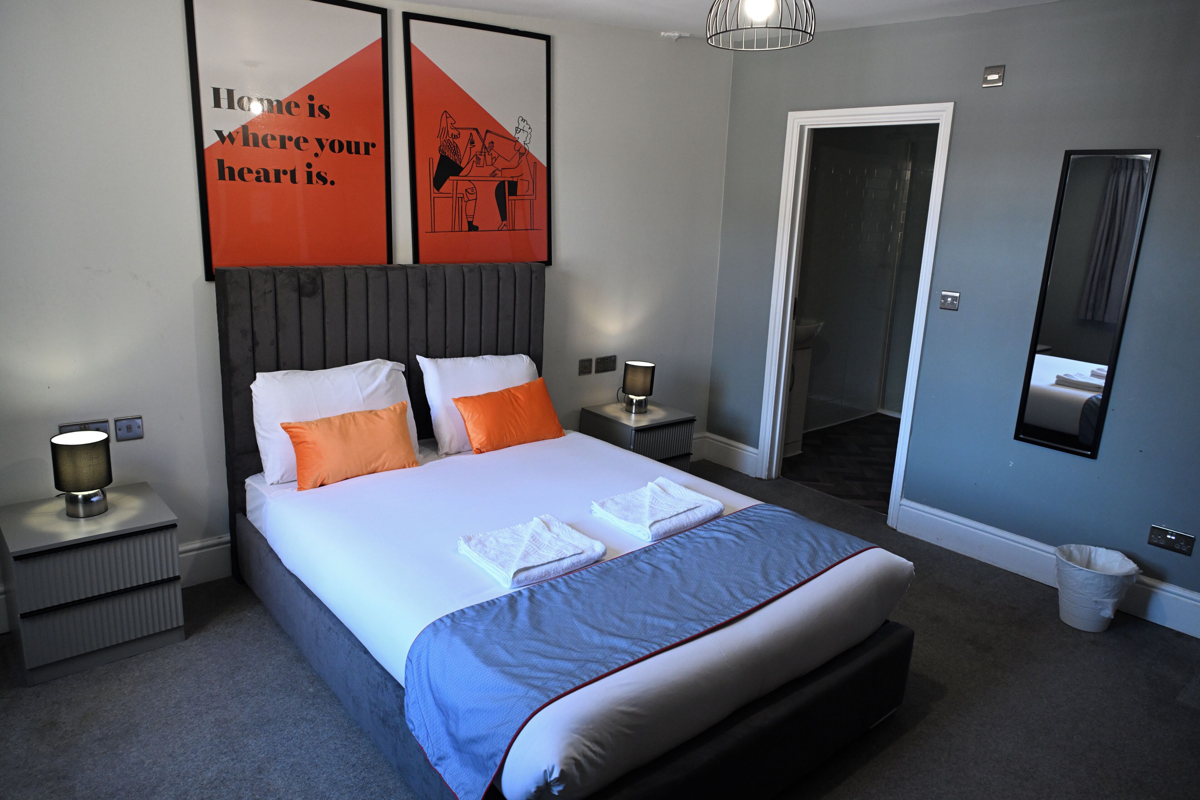 A neatly made bed with white and orange pillows, a grey headboard, and a blue bedspread. There are two bedside tables with lamps, a wall-mounted mirror, and a framed quote above the bed.
