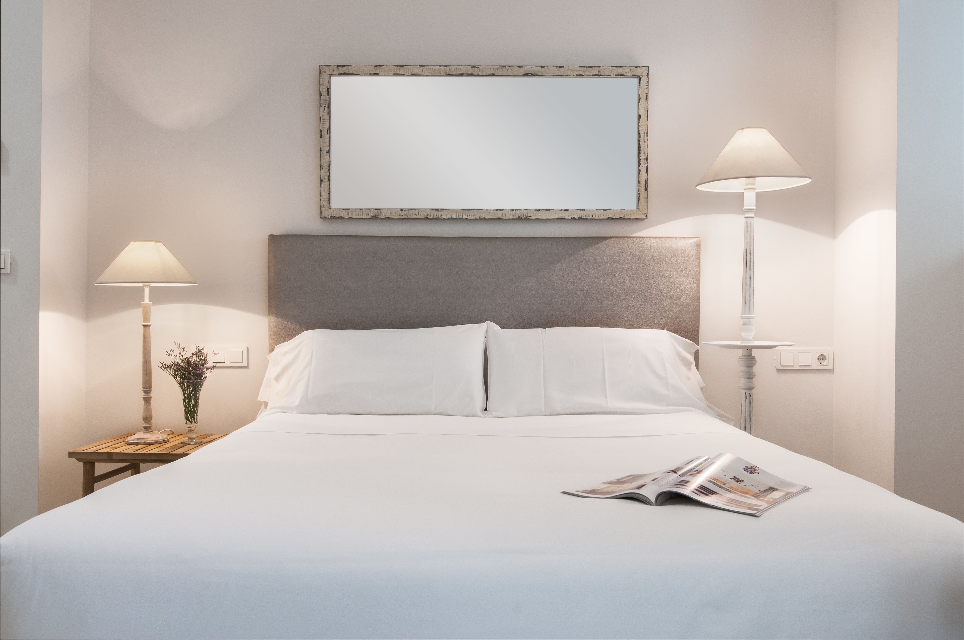 A neatly made bed with white linens, two bedside lamps, a framed mirror above the bed, and a magazine on the bed.