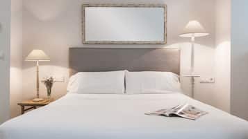 Una cama bien hecha con ropa de cama blanca, dos lámparas de noche, un espejo enmarcado sobre la cama y una revista sobre la cama.