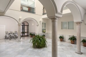 A spacious indoor area with a high ceiling, white columns, and a tiled floor. There are potted plants and bicycles visible. - Suites Sevilla Plaza Apartamentos (Seville)