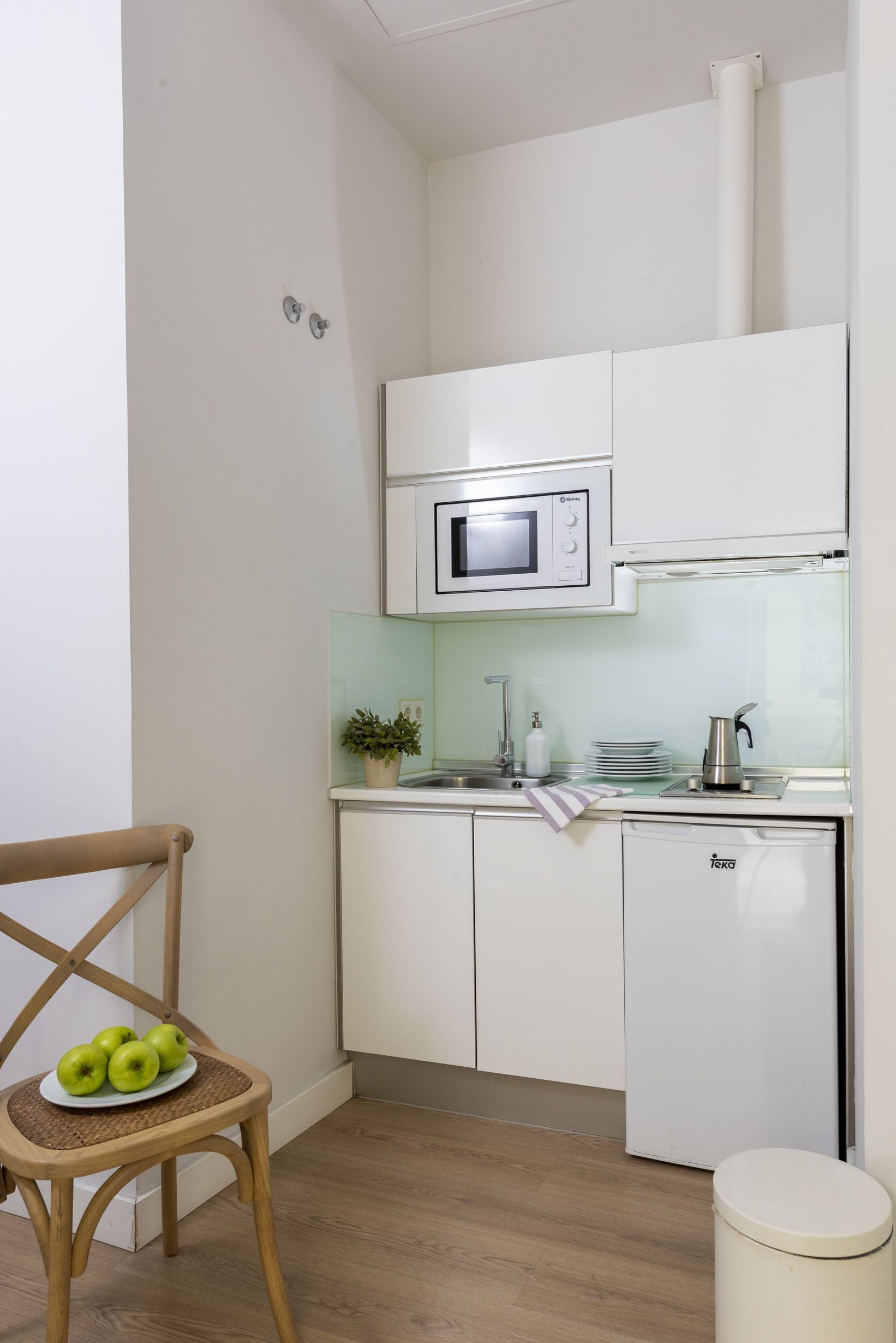 A compact kitchen with white cabinets, a microwave, a sink, and a small refrigerator. There is a wooden chair with a plate of apples and a potted plant on the counter.